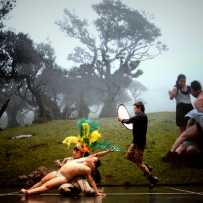 Au premier plan, 5 personnes se trouvent sur une scène de théâtre. Au centre de la photo, une personne est assise par terre, sur ses genoux. Elle porte un chapeau avec des plumes jaunes, vertes et oranges. Dans sa main droite, elle tient un stylo. Avec son bras gauche, elle tient une autre personne, allongée. La personne allongée est en culotte. Elle a des traces de stylo sur le corps. Sur la scène, au centre de la photo, il y a aussi une personne, debout qui tient un objet rond blanc et noir. En arrière plan, sur le fond de la scène, une photo de clairière est projetée. A gauche de la photo projetée, il y a des personnes en mouvement.