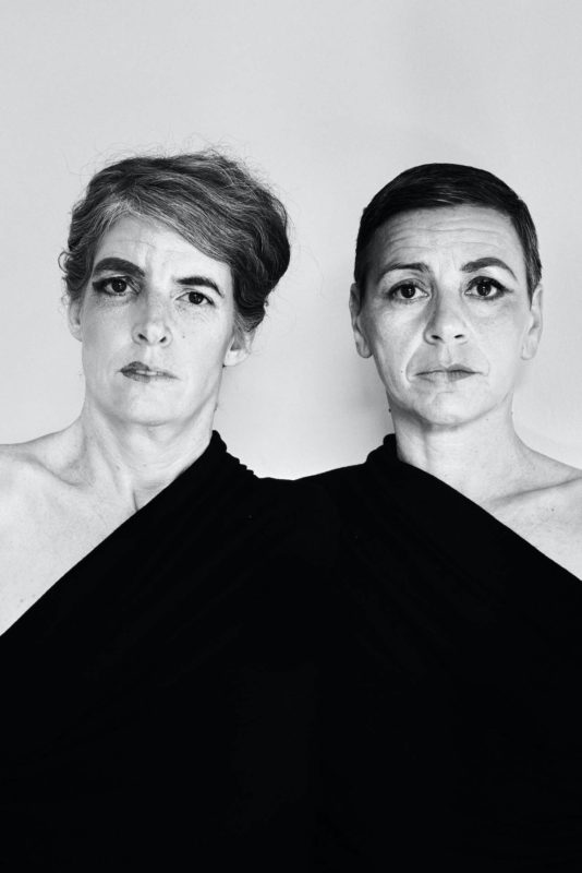 Portrait de deux femmes côte à côte: à gauche: Maud Leibundgut et à droite: Caroline de Cornière. Elles fixent l'objectif. Leurs expressions ont neutres. On voit leurs visages et leurs bustes. Elles sont toutes deux enveloppées dans un tissu noir qui couvre leurs bustes respectifs et semble les attacher l'une à l'autre. On ne voit pas leurs bras. Le tissu noir semble les unir pour faire d'elles une seule et même personne. Elles sont toutes deux maquillées seulement d'un côté.