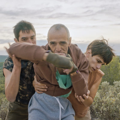 Trois hommes sont face à l'objectif. Au centre: Kamel Messelleka, ancien boxeur. Il fixe l'objectif et fait un mouvement de boxe. Derrière lui, à sa gauche et à sa droite, deux danseurs le tiennent et accompagnent ses mouvements. En arrière plan, un paysage de montagne avec beaucoup d'arbres.