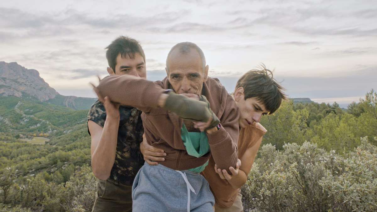 Trois hommes sont face à l'objectif. Au centre: Kamel Messelleka, ancien boxeur. Il fixe l'objectif et fait un mouvement de boxe. Derrière lui, à sa gauche et à sa droite, deux danseurs le tiennent et accompagnent ses mouvements. En arrière plan, un paysage de montagne avec beaucoup d'arbres.